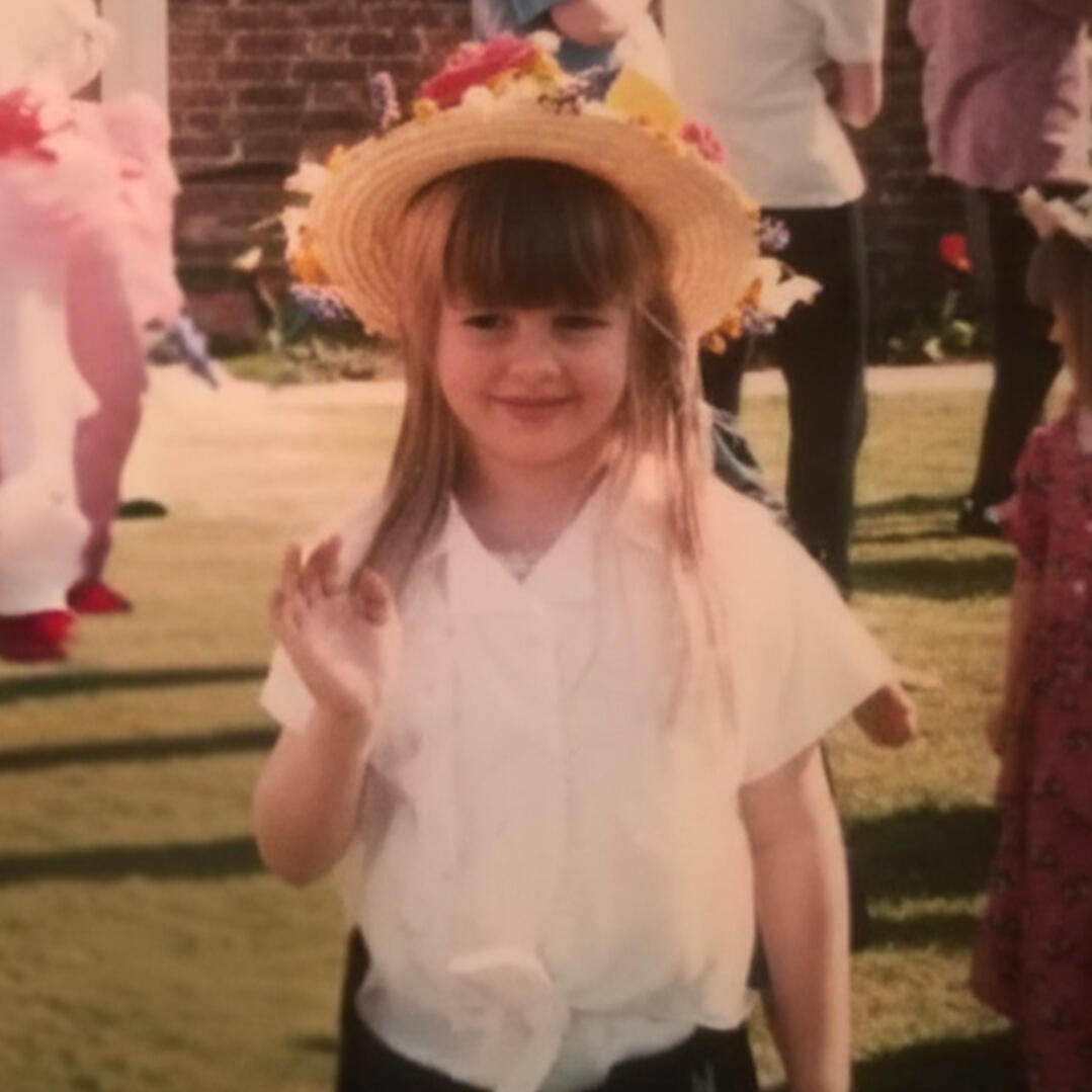 Little me Picture of Katie as a small child, wearing a flowery sunhat and waving to the camera without making eye contact.